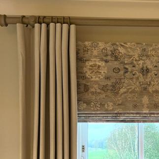 Layered roman blind in muted tones with floor length neutral curtains on a chunky wooden pole