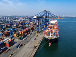 Aerial view of a busy Indian cargo port during daytime, featuring rows of colorful shipping containers, large blue cargo cranes, and a massive container ship docked beside the terminal under a clear blue sky.