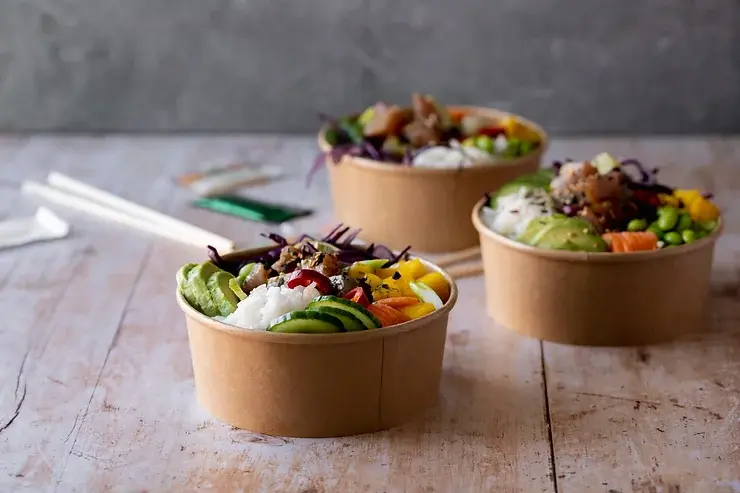 Three brown paper bowls filled with colorful poke bowls containing rice, avocado, mango, salmon, cucumbers, and other fresh ingredients, set on a rustic wooden table with chopsticks and sauce packets nearby.