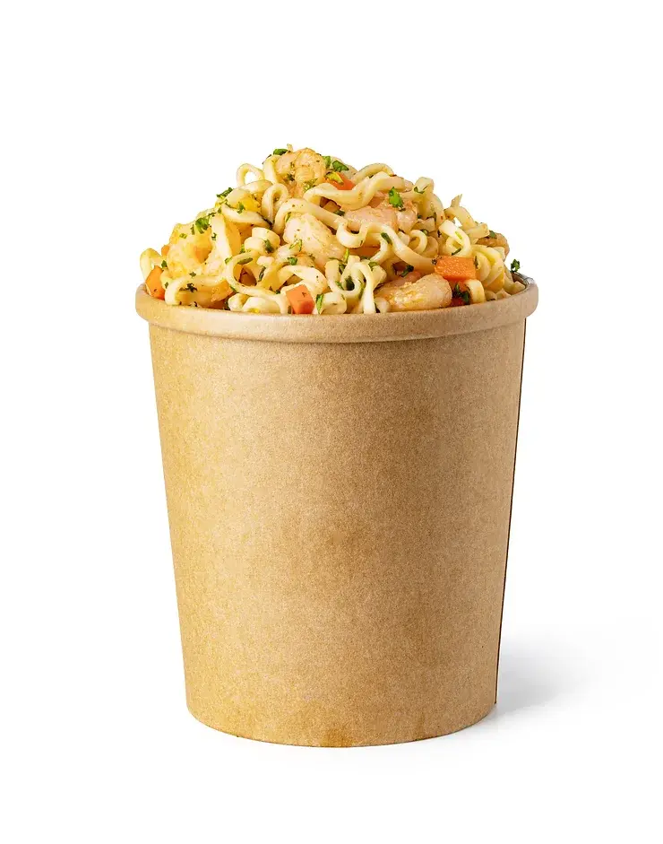 A brown paper container filled with instant noodles, garnished with shrimp, vegetables, and herbs, placed against a white background.