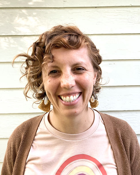 Photo of a light-skinned female with short, light brown curly hair and freckles, wearing earrings with light gold tassles, a brown sweater, and a pink shirt, against a white wood background