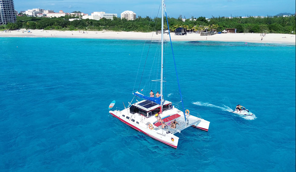 A red-and-white Pura Vida SXM catamaran glides through clear turquoise water as guests relax on deck, with a jet ski riding nearby and a tropical Sint Maarten beach in the background.