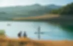 Paysage ensoleillé du lac de Saint-Cassien en Provence, eau turquoise calme avec paddle, kayak au loin et couple de randonneurs sur la berge, pins parasols et collines brumeuses, style photo magazine voyage — Le Relais Impérial