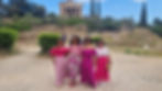 Four women in vibrant pink outfits pose together in front of an ancient Greek temple. The setting is lush and historic.