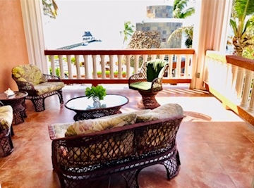 Wicker furniture with floral cushions on a sunlit balcony overlooking the ocean. Green plants and palm trees create a relaxed, tropical vibe. Belize Property Amenities