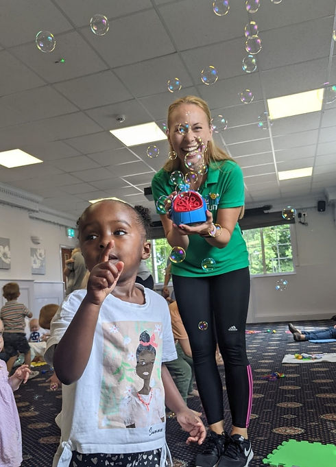 Zoé holding a bubble machine and smiling as a young child tries to pop the multiple bubbles