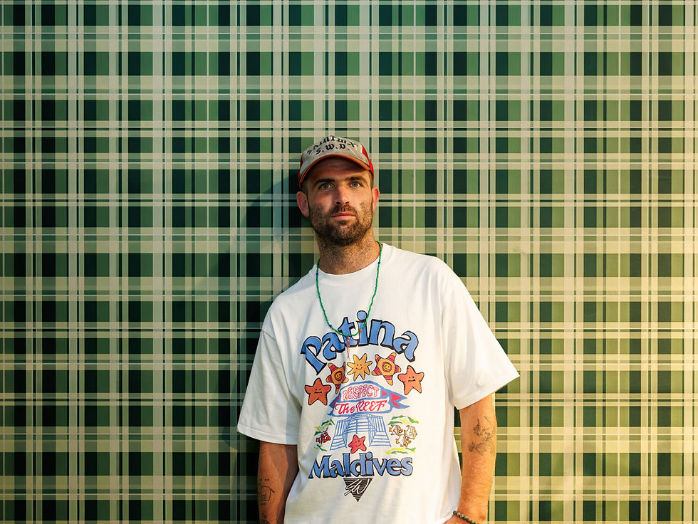 Bearded man in cap and graphic T-shirt stands against green plaid wall, looking off-camera.