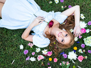 Woman in a light blue dress lies on grass, surrounded by colorful flowers. She holds a red rose, smiling joyfully under a clear sky.
