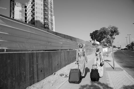 תצלום שחור־לבן של שני אנשים הולכים עם מזוודות על מדרכה עירונית, מולם קיר עץ גבוה ובניינים ברקע.