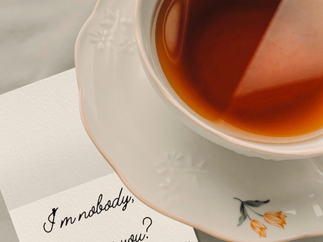 A tea cup, filled sits on a table with a note tucked underneath. On it reads "I'm nobody, who are you? --Emily Dickenson"