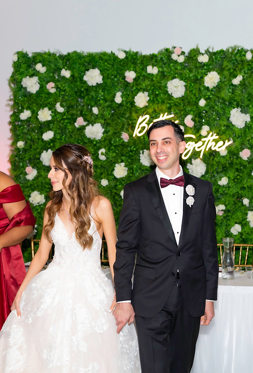 Bride and groom smiling inside banquet hall with guests in background.