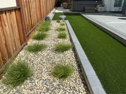 Modern side yard with synthetic grass, belgard midnight paver edging, and ornamental grasses in a light-colored river rock bed in Bethel Island.