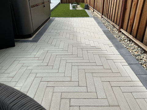 Close-up of light grey Belgard plank pavers in a herringbone pattern with a charcoal border next to a wood fence and turf.