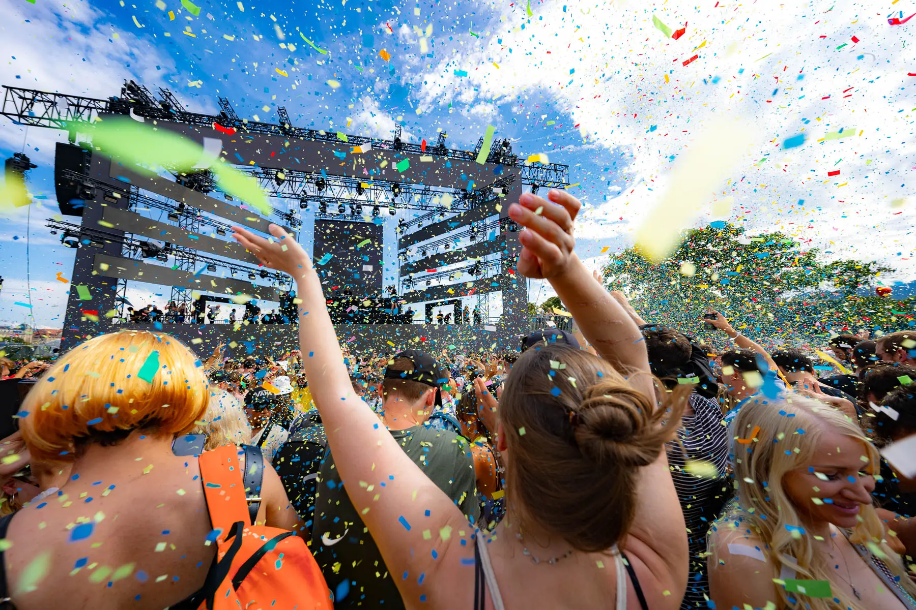 Menschenmenge feiert vor Bühne im Freien mit buntem Konfetti in der Luft, jubelnde Arme.