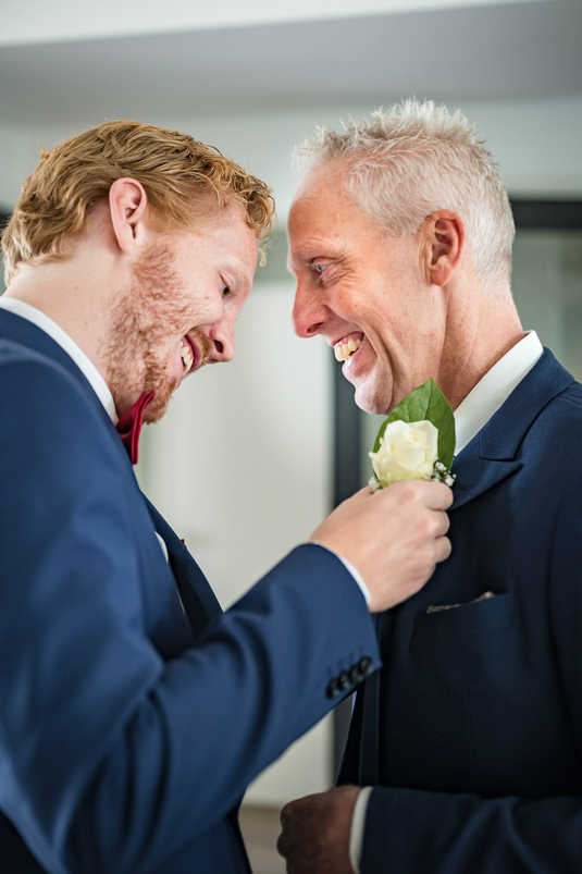 Beste vriend van bruidegom en diens vader lachen naar elkaar terwijl zij de corsages opspelden.