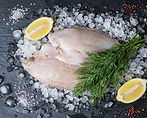 Two raw lemon sole fillets layered on a dark slate board with lemon wedges and peppercorns.