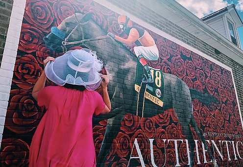 Woman in a pink dress wearing a fancy Derby hat, looking at a mural of Kentucky Derby winner Authentic