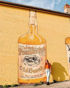 Woman standing in front of a huge mural showing a bottle of John Poindexter Old Bourbon