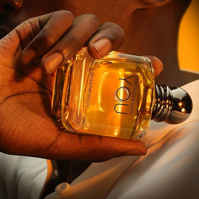 Mão segura frasco de perfume "Stronger With You" da Emporio Armani, líquido dourado, fundo laranja suave, transmitindo elegância.
