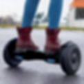Pessoa de jeans e botas vermelhas em Hoverboard Skate Elétrico 10" da Brinovar com luzes azuis, sobre asfalto molhado ao ar livre.