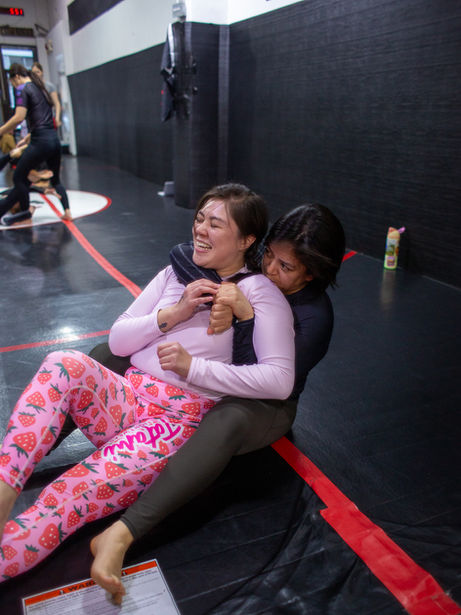 Two women laughing while grappling on a black and red wrestling mat About Us.