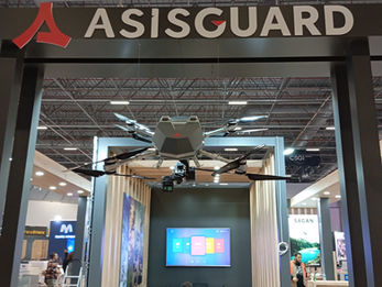 ASISGUARD için üretilen, mat siyah çatı alın tabelası, tavan askılı drone sergileme alanı ve ahşap konsept toplantı bölmesi içeren fuar standı genel görünümü.