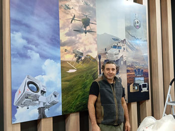 ASISGUARD fuar standının iç mekân ahşap dikey çıtalı duvarı üzerine uygulanan çoklu ürün ve drone görselli dijital baskı panel giydirmesi.