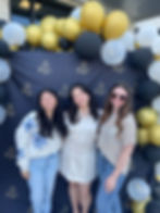 Three women posing in front of a black and gold balloon arch.