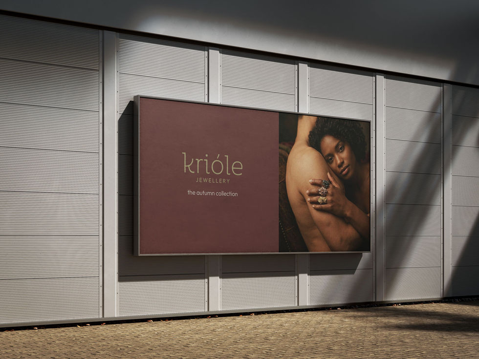 
A horizontal billboard for "Krióle Jewellery" is mounted on a grey slatted wall above a cobblestone path. The left side of the advertisement has a deep burgundy background featuring the brand name and "the autumn collection" in gold text. The right side shows a photograph of a woman with curly hair leaning against a man's shoulder, showcasing three large, ornate flower-shaped rings on her fingers.