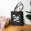 Thumbnail: A black cotton canvas tote bag with a flying superhero dog in graphic stands on the floor