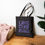 Thumbnail: Black Cotton canvas bag with tagline, like this bag in lavender color, stands on the floor