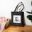 Thumbnail: Black cotton canvas tote bag with a chill vibe cat relaxing on summer beach chair stands on the floor.