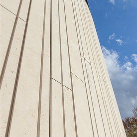 Looking up a soaring architectural pillar clad in vertical Khaki Limestone panels of different widths with a finely textured, linear surface under a clear sky.