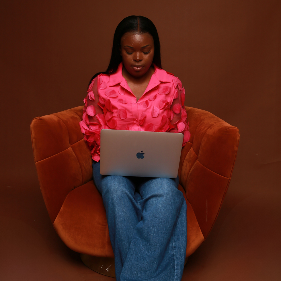Black woman wearing a pink blouse, sitting in a brown chair, working on a laptop, demonstrating focus and self-care through setting workplace boundaries.