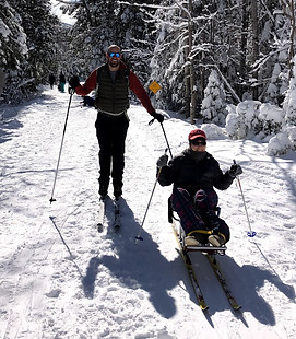 A standing skier poses next to a seated skier on a snow covered woodland trail.