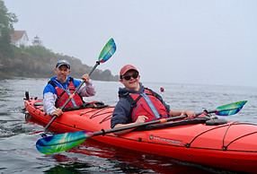 Two white men in a two person red sea kayak. They are on the water and are wearing red life jackets and holding green kayak paddles.