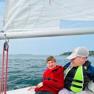 A boy in a red life jacket sits next to a man in a green life jacket. There is a sail above them and water behind them.