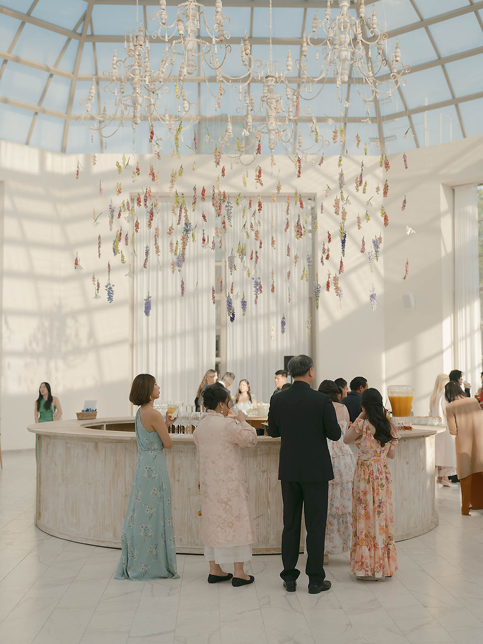 Guests gathered at the conservatory bar beneath cascading hanging florals and chandeliers, illuminated by late afternoon light.