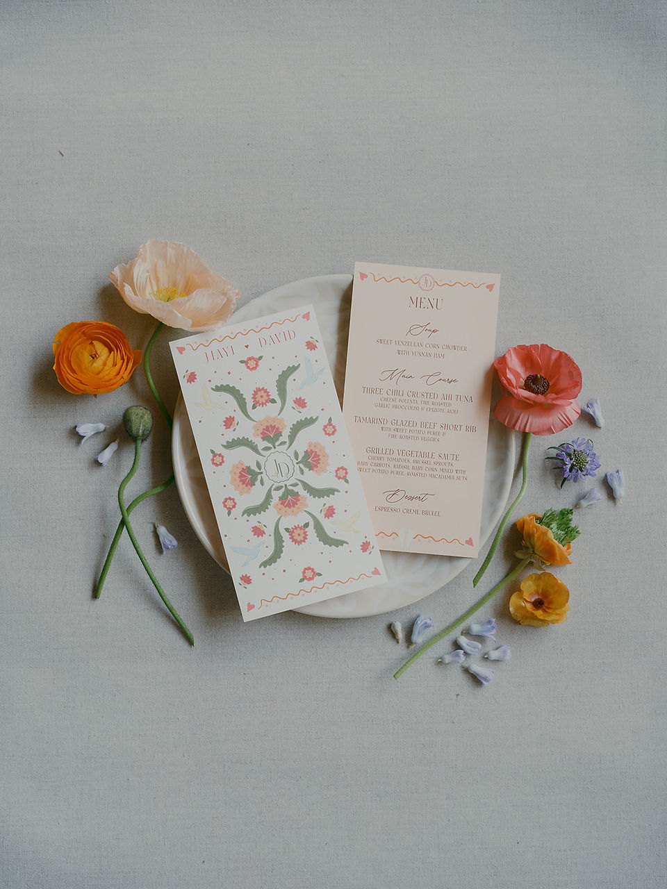 Flat lay of hand-painted wedding stationery and florals on pale blue linen — featuring ranunculus and calligraphy menus.