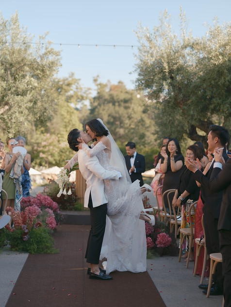 Bride and groom sharing their first kiss surrounded by guests clapping along the floral-lined ceremony aisle at Monserate Winery.