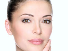 Woman with tied-back hair touches her face, gazing softly. Neutral background, clear complexion, serene expression.