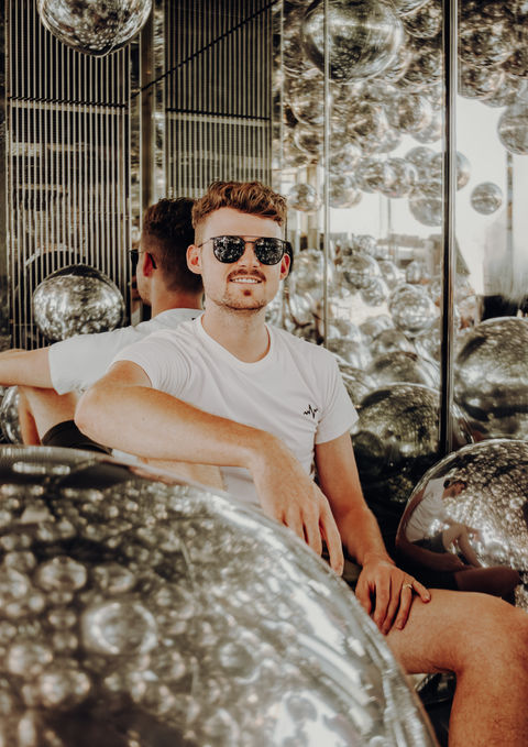 Ein Mann mit Sonnenbrille sitzt in einem verspiegelten Raum in einem Hochhaus mit Bällen in Chrom-Optik.