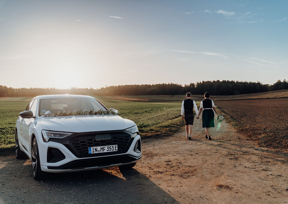 Bayerisches Brautpaar in blau-weißer Tracht von hinten auf einem Feldweg weggehend während eines Sonnenuntergangs mit Brautauto, ein Audi Q8 e-tron