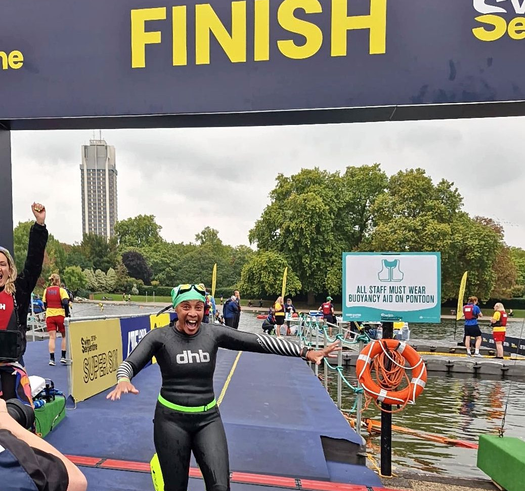 Smiling swimmer in black wetsuit celebrates at FINISH on pontoon by Serpentine Lido.