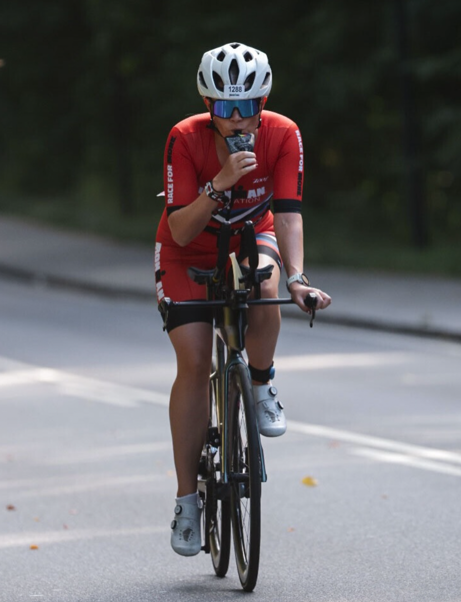 Triathlete in red trisuit with white helmet fuelling on a gel while riding her bike.