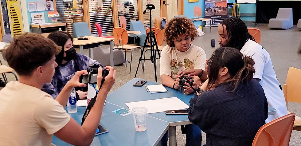 Five people in a colorful room with cameras and papers on a table. One is explaining camera settings. Relaxed, collaborative mood. learning and mentorship.