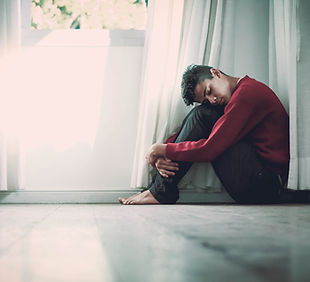 Image of a young man sitting by a window and holding his knees