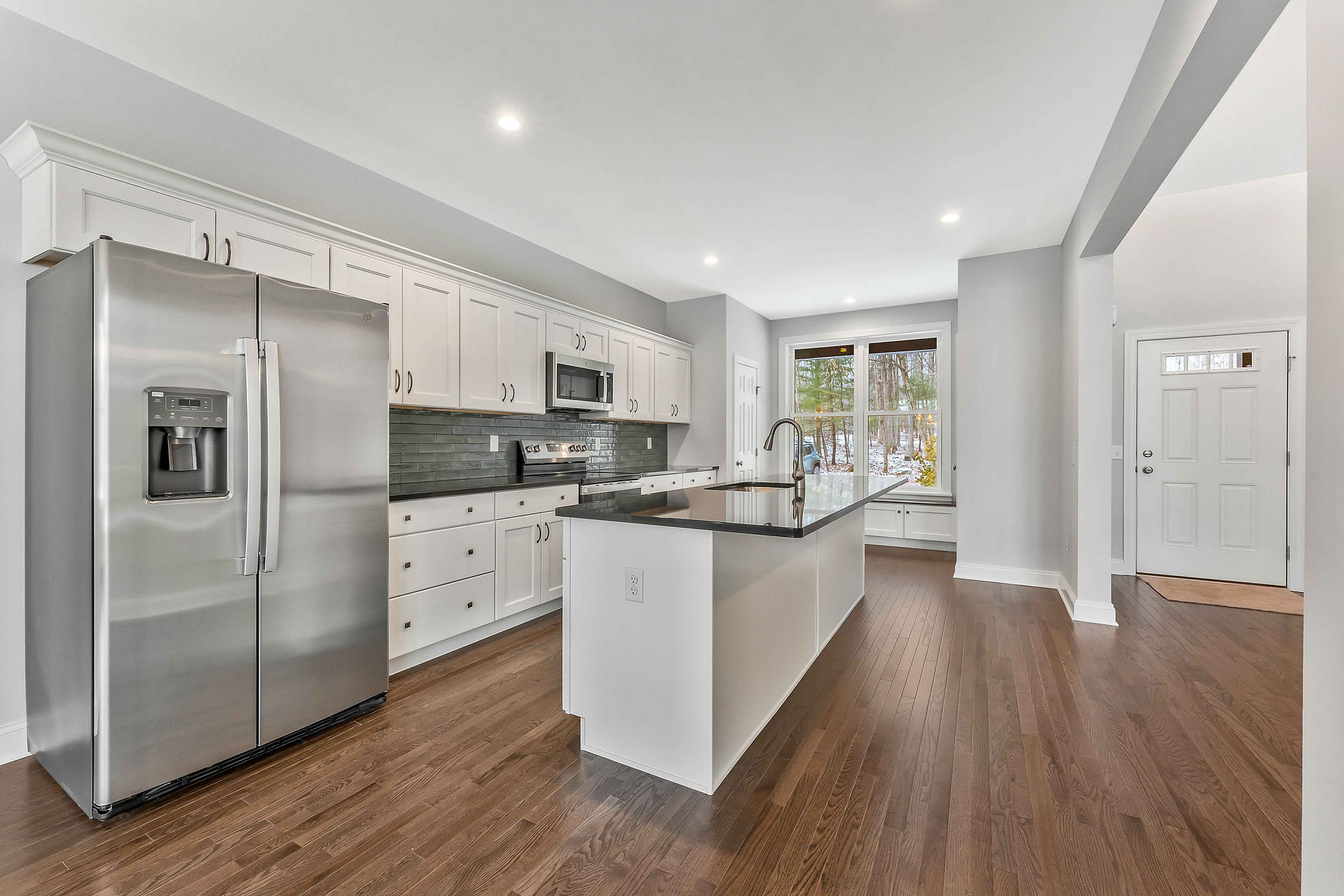 Finished kitchen with a long white island and dark countertop, stainless steel refrigerator and range, white shaker cabinets, green tile backsplash, recessed lighting, hardwood floors, and a window seat near the entry door.