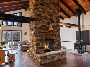 Furnished living room area with a stone fireplace in the center. The room has high, wooden-beamed ceilings and large windows that provide views of trees and a lake. A tan sofa is visible in the background, and a wooden table with some items on it is to the left. On the right side, a wood stove is seen, as well as a small metal container.