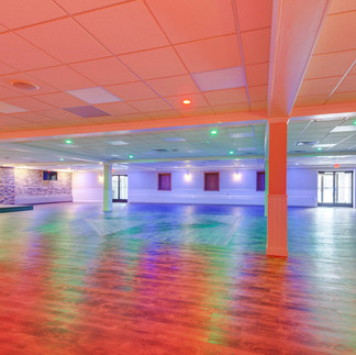 Large event hall with wood-style flooring, stone accent walls, and white support columns. The drop ceiling has recessed lights with colorful accent lighting in red, green, and purple, casting vibrant reflections across the floor and walls. Double glass doors provide entry at the far end of the room.